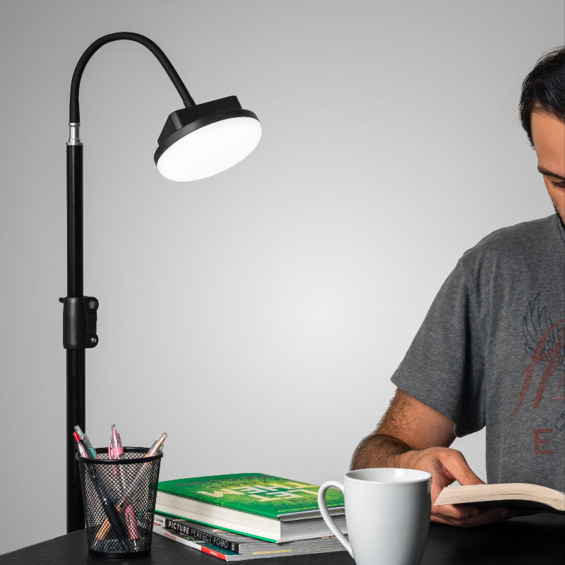 Person reading a book under  adjustable desk lamp with a cup and books on the table.